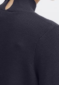 Pull en tricot navy foncé avec un col haut et un dos ouvert. Présente une texture côtelée avec une coupe ajustée. Pas de matériel visible.