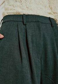 Tmavozelené skladané nohavice vyrobené z textúrovaného materiálu, s pásom s pútkami na opasok a bočnou vreckom.