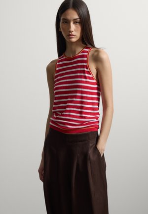 Femme aux longs cheveux foncés portant un haut sans manches rayé rouge et blanc et un pantalon marron foncé taille haute, debout devant un fond uni.