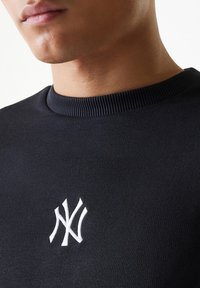 Sudadera negra con cuello acanalado y un prominente logo bordado en blanco de los New York Yankees en el pecho.