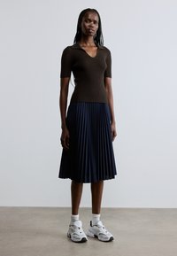 Top marrón de manga corta con textura acanalada y escote en V, combinado con una falda plisada azul marino. Zapatillas blancas con detalles en negro y cordones visibles.