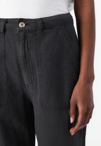 Un pantalon en lin noir présente une finition texturée, une fermeture à bouton et des poches latérales, avec une coupe décontractée qui met en valeur le drapé du tissu.