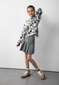 Biała bluza z czarnymi wzorami graficznymi, szara spódnica plisowana i beżowe sneakersy. Długie włosy swobodnie opadające, ręce uniesione w zabawny sposób.