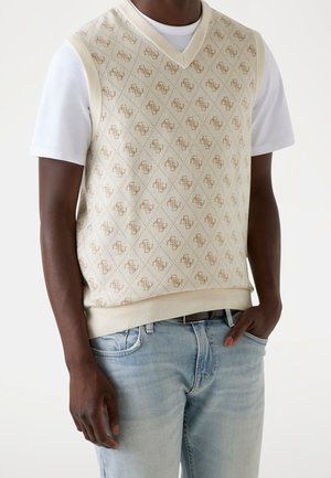 Homme portant un gilet en tricot crème avec un motif monogramme beige sur un t-shirt blanc et un jean bleu clair, main dans la poche, sur fond uni.