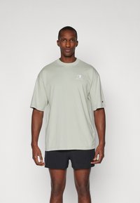 Lys grøn bomulds T-shirt med korte ærmer og rund hals, der har et lille logo på brystet. Bærende med mørke shorts.