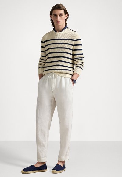 Pull en tricot crème avec des rayures bleu marine, pantalon en lin blanc ample et espadrilles bleu marine. Le pull présente un col rond et des détails de boutons sur le côté.