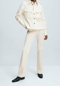 Chemise boutonnée couleur crème avec deux poches poitrine, associée à un pantalon large assorti et des bottines à talons noires, sur un fond neutre.