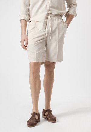 Persona que lleva pantalones cortos beige con cordón, una camisa beige de manga larga remangada y sandalias cerradas de ante marrón.