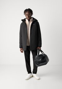 Veste rembourrée noire avec une capuche, polaire crème à fermeture éclair en dessous, pantalon noir et baskets blanches. Tenant un sac de voyage en cuir noir.