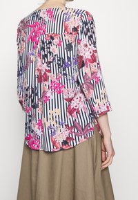 Kobieta w paskowanej bluzce z różowymi i fioletowymi wzorami kwiatowymi oraz jasnobrązowej spódnicy, pokazana z tyłu z jedną ręką luźno opadającą.