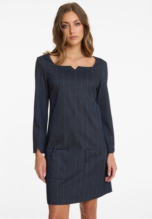 Femme portant une robe bleu marine à rayures verticales, mi-longue, à manches longues, avec un décolleté carré et des poches plaquées à l'avant, sur fond blanc.