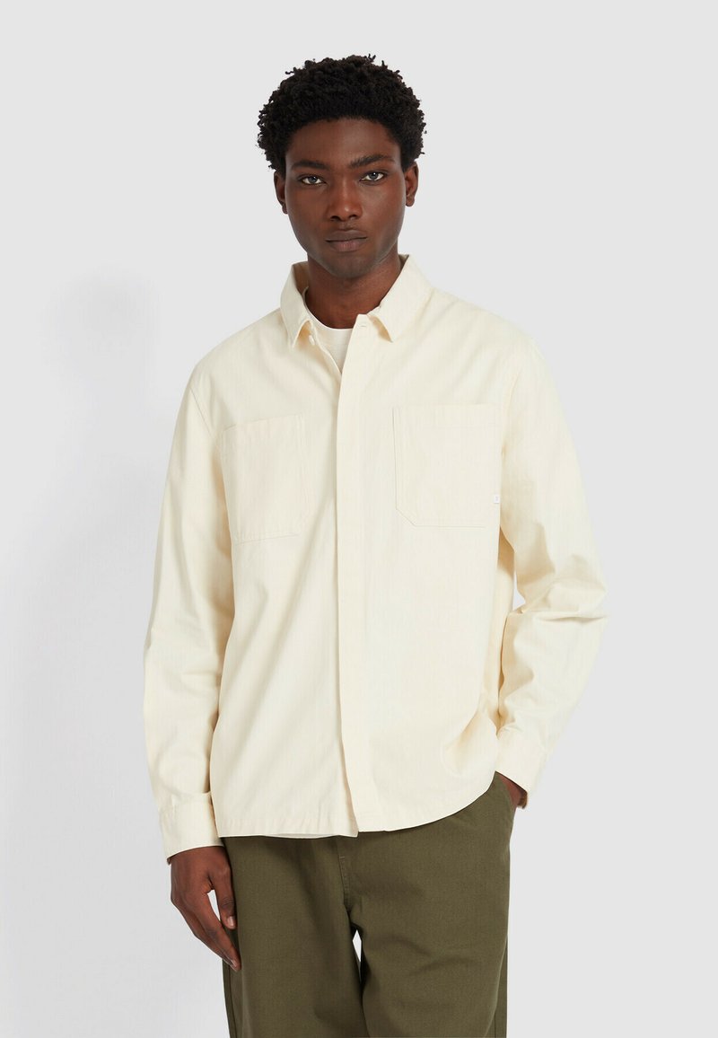 Mężczyzna ubrany w kremową koszulę na guziki z kieszeniami na piersi oraz oliwkowe spodnie, stojący z jedną ręką w kieszeni na tle jednolitym.