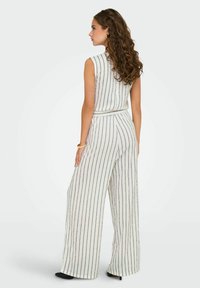 Jumpsuit bianco a righe verticali con design a gamba larga, top senza maniche, tessuto texturizzato e vita aderente, abbinato a tacchi neri.