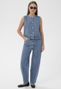 Frau trägt eine ärmellose, geknöpfte Jeansweste, passende hochgeschnittene, weite Jeans, schwarze Ballerinas und schwarze ovale Sonnenbrillen und posiert.