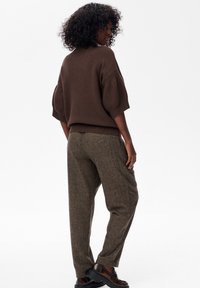 Vrouw met krullend haar, gekleed in een bruine sweater met korte mouwen, grijs-bruine op maat gemaakte broek en donkerbruine instappers, staand tegen een witte achtergrond.