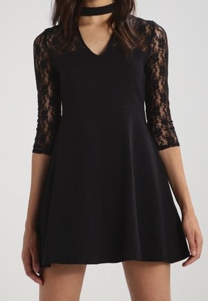 Femme portant une robe noire avec des manches trois-quarts en dentelle transparente et un col ras du cou, debout devant un fond uni.