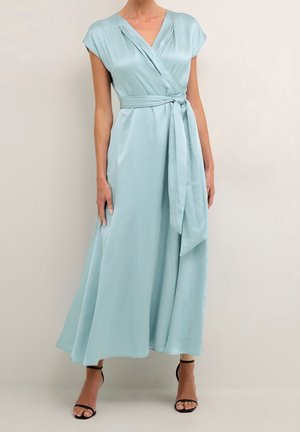 Kobieta ubrana w jasnoniebieską sukienkę kopertową z krótkimi rękawami i paskiem w talii, w połączeniu z czarnymi sandałami na wysokim obcasie z paskami.