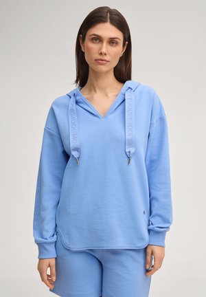 Vrouw draagt een lichtblauwe hoodie met brede trekkoorden en bijpassende korte broek, staand tegen een effen lichte achtergrond.