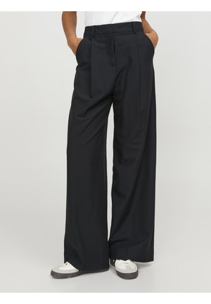 Personne portant un pantalon noir à jambes larges et des baskets blanches, les mains dans les poches, debout devant un fond clair et uni.