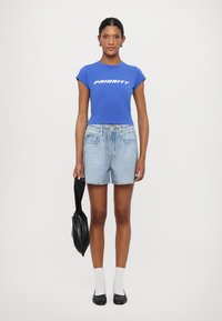 T-shirt cropped blu con il testo "PRIORITY", pantaloni shorts in denim blu chiaro, calzini bianchi alla caviglia, ballerine nere e una borsa nera. Un outfit semplice e informale.