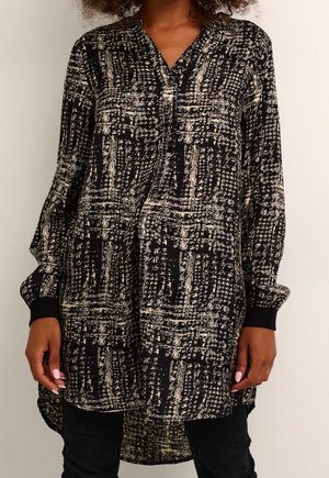 Schwarz-beige gemusterte Tunika mit langen Ärmeln, V-Ausschnitt mit Knöpfen und gekrempelten Ärmeln, getragen über schwarzen Hosen.