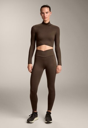 Eine Frau trägt ein passendes braunes Langarm-Crop-Top und hoch sitzende Leggings mit schwarzen Sportschuhen und steht vor einem neutralen Hintergrund.