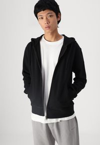 Sudadera negra con cremallera, capucha con cordones, bolsillos frontales y puños acanalados, superpuesta sobre una camiseta blanca, combinada con pantalones de chándal grises.