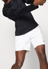 Miestenský športový outfit pozostávajúci z čierneho trička s dlhým rukávom a bielych šortiek s jemným logom, zdôrazňujúci ľahký materiál a športový strih.