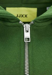 Sudadera con cremallera verde que presenta un cuello alto, cremallera plateada y textura acanalada. Etiqueta con "JJXX" en amarillo ubicada dentro del cuello.