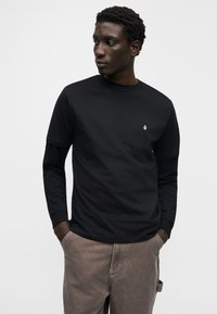 Μαύρο μακρυμάνικο μπλουζάκι από βαμβάκι, με στρογγυλή λαιμόκοψη και μικρό λευκό λογότυπο στο στήθος. Φορεμένο με καφέ παντελόνια.