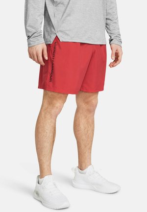 Mężczyzna ubrany w czerwone spodenki Under Armour, szarą koszulkę z długim rękawem i białe buty sportowe, stojący na białym tle.