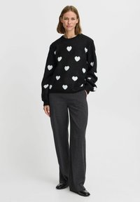 Schwarzer Pullover mit weißem Herzmuster, weicher Textur, runder Ausschnitt und langen Ärmeln, kombiniert mit grauen, weit geschnittenen Hosen und schwarzen Schuhen.
