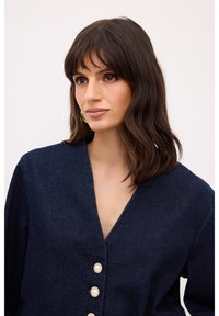 Chaqueta de mezclilla azul marino con escote en V, que presenta cuatro botones decorativos con centros de perla y un corte relajado.