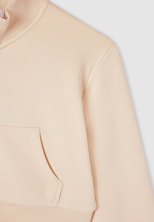 Felpa beige chiaro con collo alto, polsini a costine e tasca laterale. Tessuto morbido con texture liscia e dettagli di cucitura discreti.