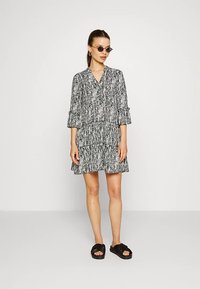 Vestido com padrão preto e branco, com decote em V, cintura franzida e saia em camadas. Mangas compridas com punhos. Modelo usando sandálias pretas e óculos de sol.