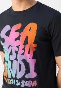 Close-up of a person wearing a black T-shirt with large, colorful gradient text reading "SEA YOURSELF AND I" and smaller text below.