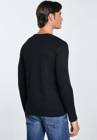 Vyras, dėvintis juodą ilgomis rankovėmis marškinius ir mėlynas džinsus, stovintis nugara ir galva šiek tiek pasukta į dešinę, žemame fone.