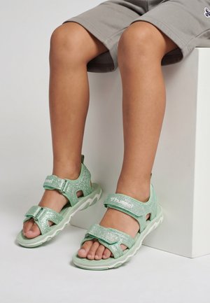 Barn iført lysegrønne glitrende sandaler med justerbare stropper, siddende med bøjede knæ på en hvid blok.