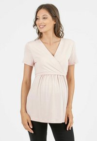 Jasnoróżowa bluzka z krótkim rękawem, dekoltem w kształcie litery V i talią empire, z funkcjonalnym fasonem kopertowym oraz miękkim, elastycznym materiałem. W połączeniu z czarnymi spodniami.