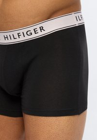 Κοντινό πλάνο ενός ατόμου που φοράει μαύρα μποξεράκια Tommy Hilfiger με λευκή ελαστική μέση με λογότυπο.