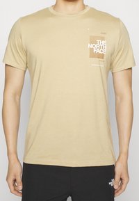 Béžové bavlněné tričko s krátkým rukávem, kulatým výstřihem a grafickým designem na přední straně, který zobrazuje "THE NORTH FACE" a čáry.