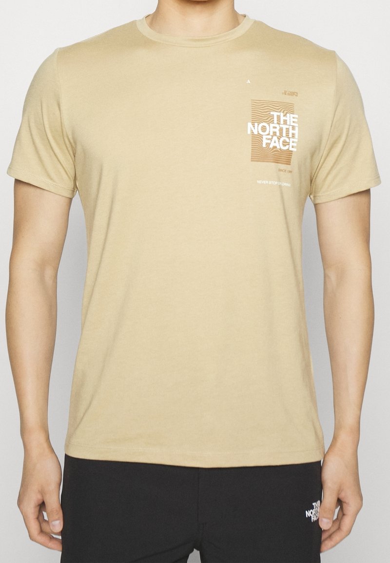 Béžové bavlněné tričko s krátkým rukávem, kulatým výstřihem a grafickým designem na přední straně, který zobrazuje "THE NORTH FACE" a čáry.