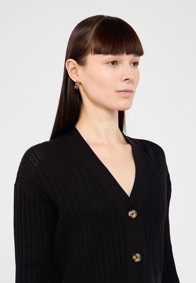 Cardigan noir côtelé avec un décolleté en V et trois grands boutons. Le tissu semble texturé et léger. Des boucles d'oreilles agrémentées d'or complètent le look.
