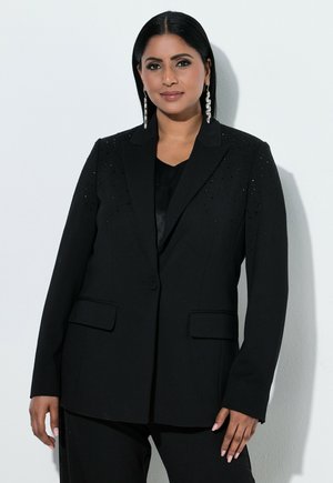 Frau mit glatten schwarzen Haaren, die einen schwarzen Blazer mit dezenten Verzierungen und langen silbernen Ohrringen trägt, steht vor hellem Hintergrund.
