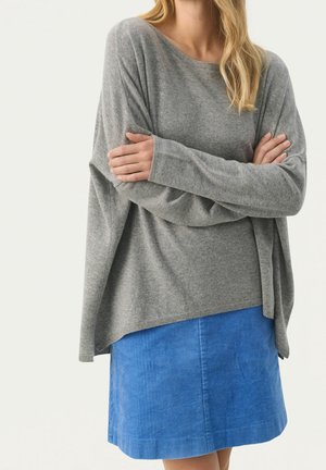 Femme croisant les bras, portant un pull gris ample et une jupe en velours côtelé bleue jusqu’aux genoux sur un fond uni.