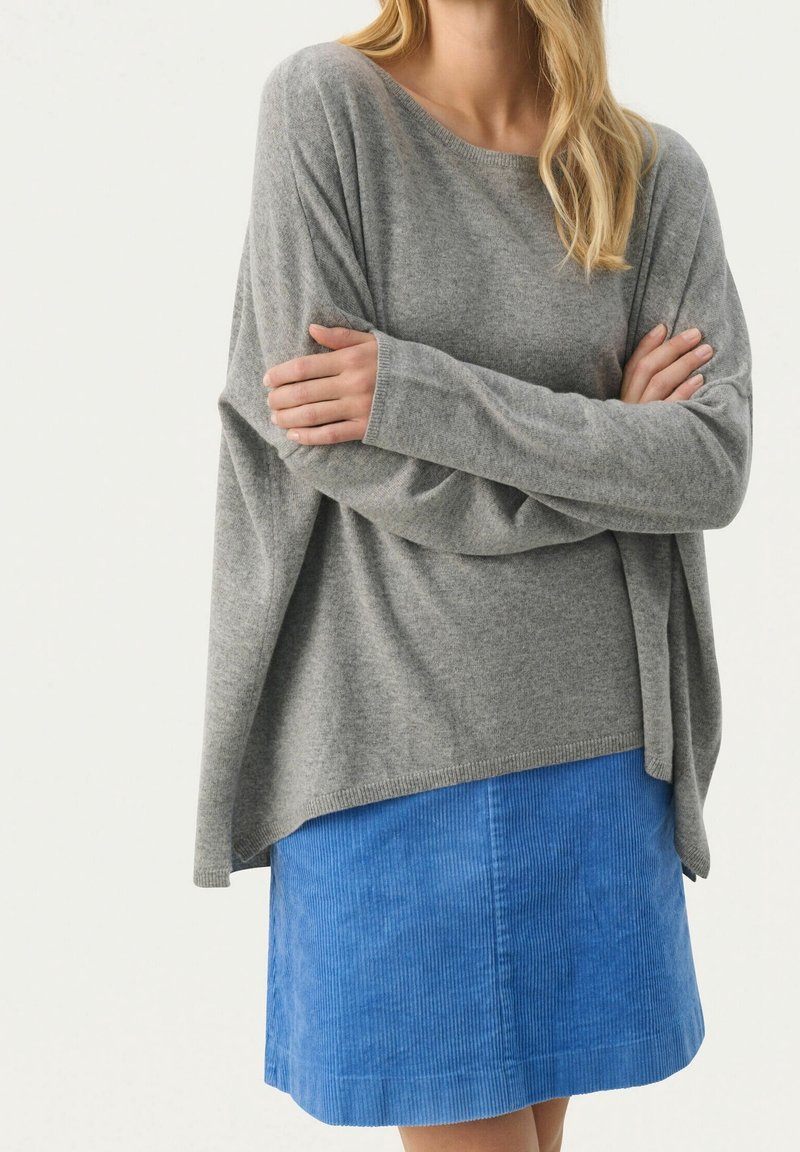 Femme croisant les bras, portant un pull gris ample et une jupe en velours côtelé bleue jusqu’aux genoux sur un fond uni.
