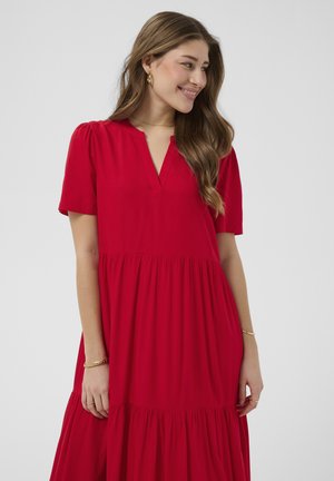 Femme aux longs cheveux bruns portant une robe rouge à manches courtes avec un col en V, souriante et regardant sur le côté sur un fond uni.