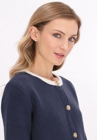Cardigan blu navy con una tessitura a rilievo, colletto bianco e bottoni dorati. Primo piano del capo, che mette in evidenza il suo design e i dettagli.