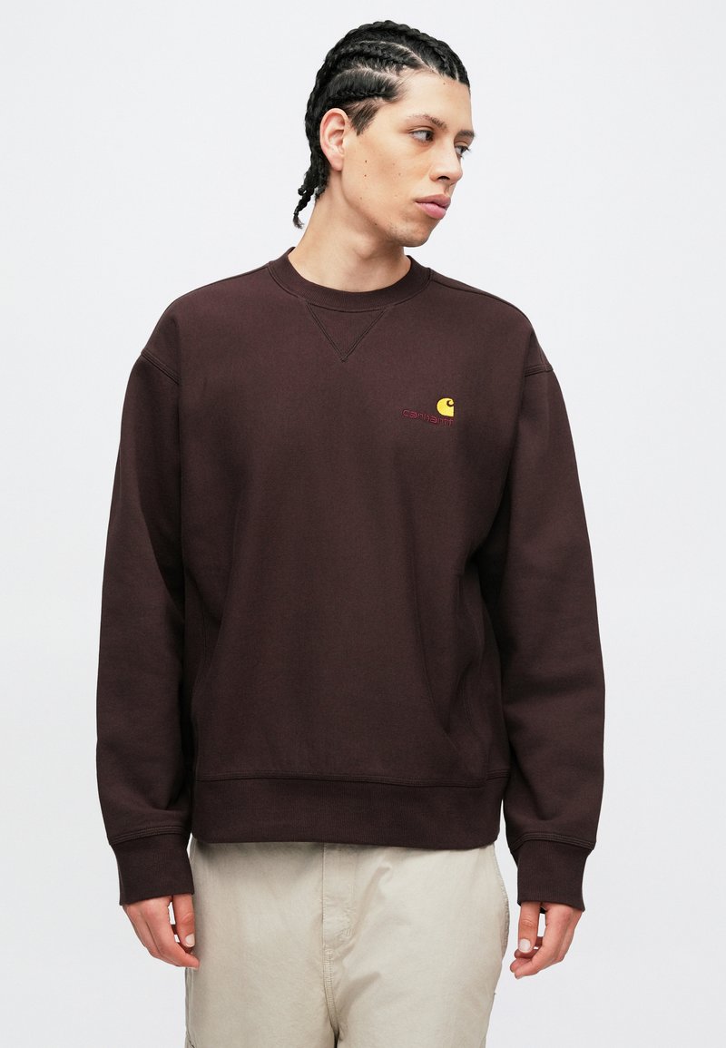 Dunkelbraunes Sweatshirt aus weichem Stoff, mit Rundhalsausschnitt und gerippten Bündchen, versehen mit einem kleinen gelb-roten Logo auf der linken Brust.