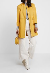 Keltainen villasekoitekotelo, jossa on pyöreä pääntie, etutaskut, yhdistetty harmaan pooloneuleen, kermafärisillä leveälahkeilla housuilla ja valkoisilla korkokengillä.